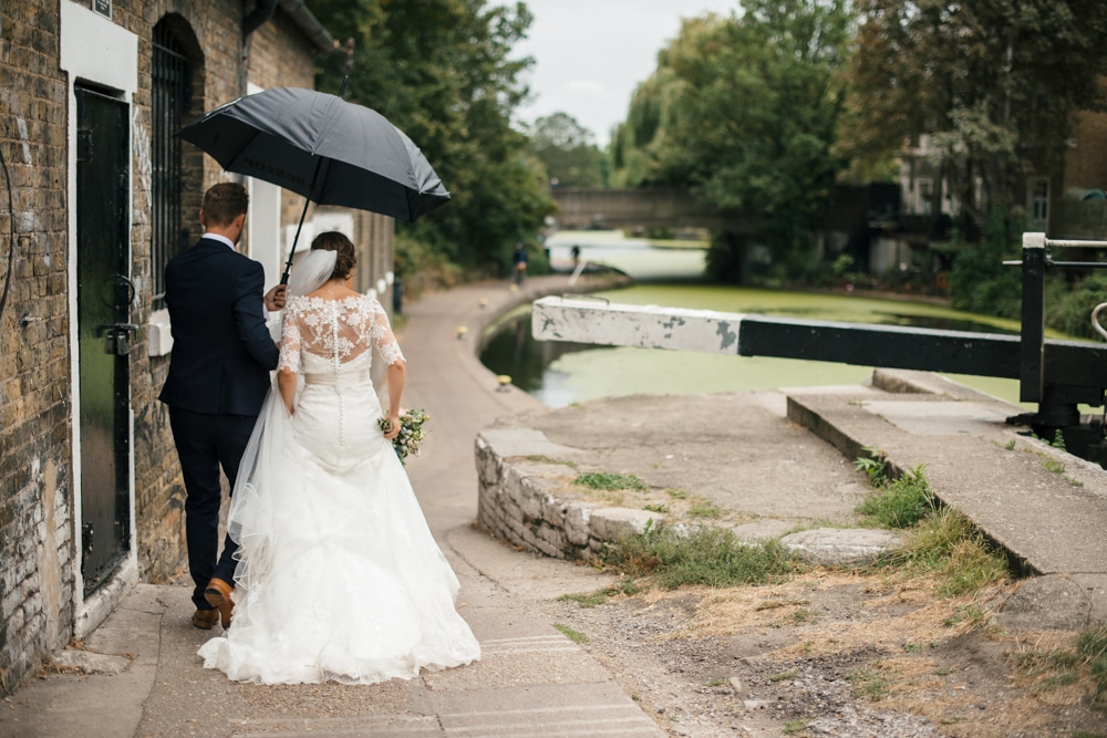 Bride and groom under umbrella walking along canal