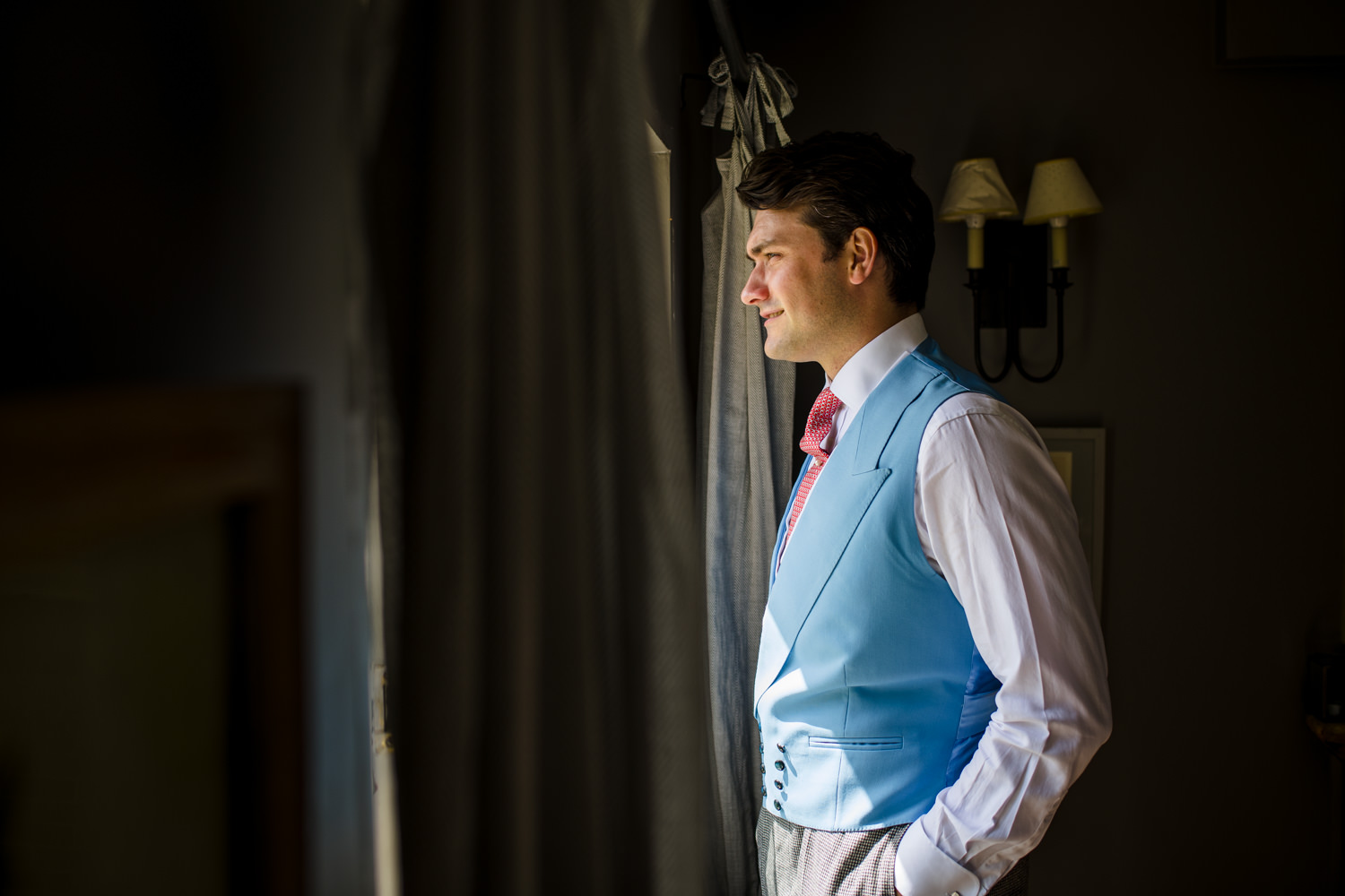 groom in pink and light blue waistcoat looking out window
